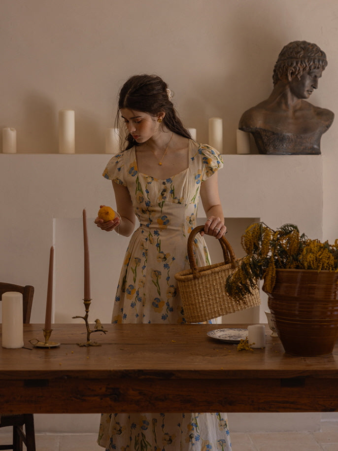 A young woman gazes softly to the side while resting her hand gently on a rustic wooden table. She wears a cream floral dress with blue and yellow blossoms, evoking a serene, vintage-inspired charm against a warm, muted backdrop with dried flowers and candle accents.
