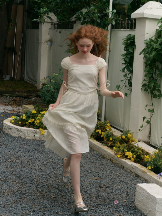 A young woman with long red hair runs joyfully on a gravel path, wearing a flowing ivory puff-sleeve dress with waist ties fluttering in the air. She is surrounded by blooming yellow flowers and white garden walls, capturing a lively and whimsical moment.