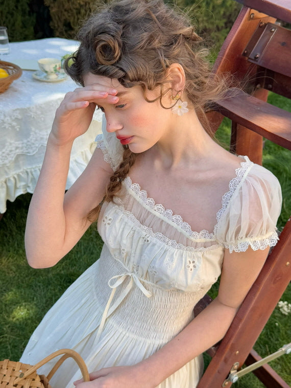A young woman in a fairytale-inspired ivory dress with puff sleeves and delicate lace trim sits on a wooden chair in a garden, gently touching her forehead, evoking a vintage cottagecore aesthetic.