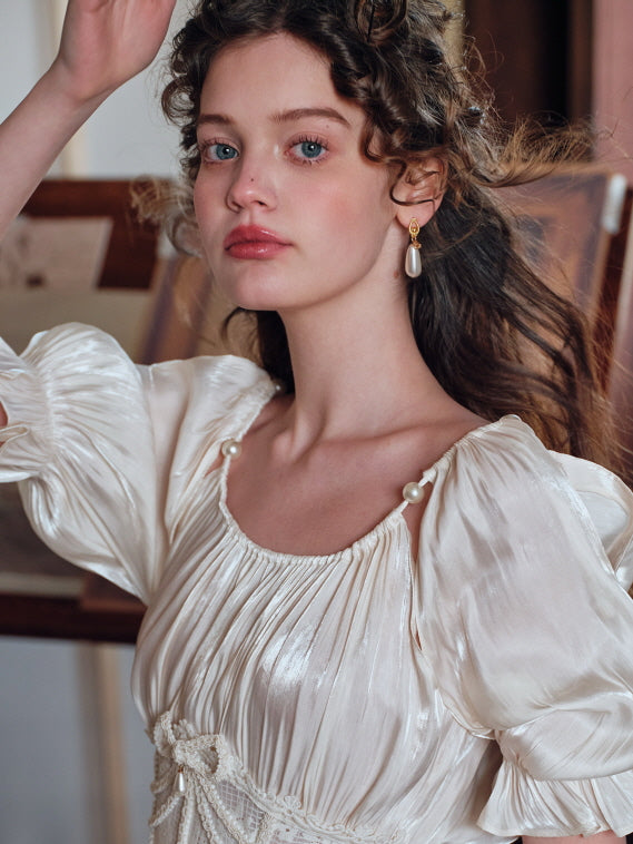 A romantic close-up of a woman wearing a vintage-inspired ivory satin camisole top with pearl shoulder accents and pleated detailing, her soft curls and pearl earrings enhancing the dreamy, fairytale aesthetic of the look.