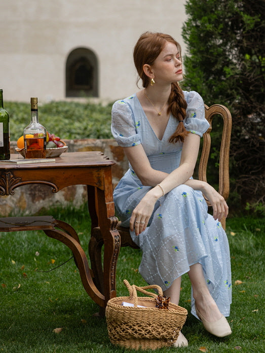 A woman in a pastel blue floral dress sits gracefully at an outdoor vintage wooden table set with fruits, a basket bag, and a bottle of wine, creating a dreamy cottagecore summer scene.