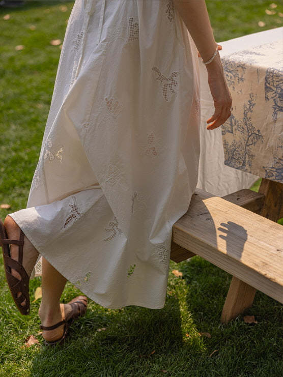 A close-up of a white embroidered dress with delicate floral and butterfly lace patterns, styled with brown leather sandals on green grass.