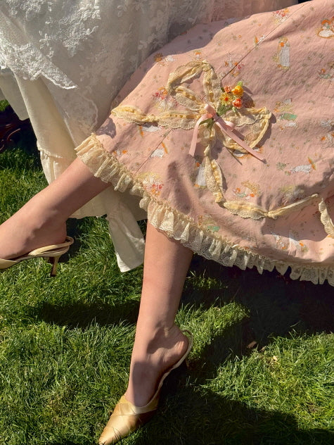 Close-up of a delicate pink floral lace dress with ruffled hem and bow accents, paired with gold vintage-style slippers in a sunlit garden setting.