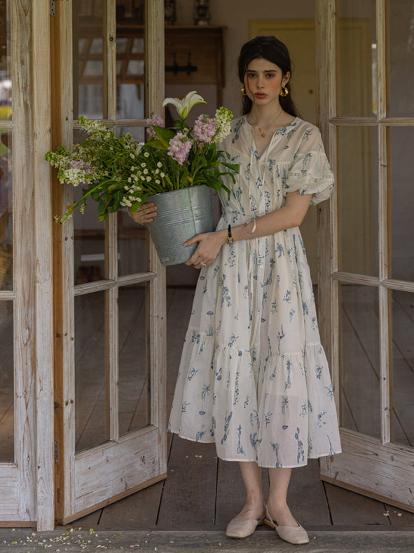 A woman in a vintage-inspired floral dress holding a metal bucket filled with pink and white flowers, standing in front of wooden French doors, creating a romantic cottagecore aesthetic.