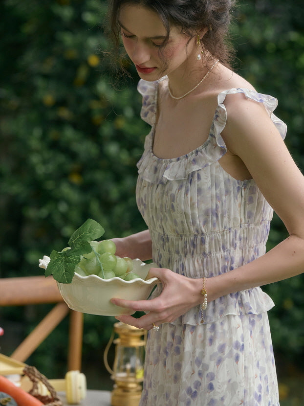 A woman in a lavender floral cottagecore dress stands holding a bowl of green grapes at a garden picnic table, creating a dreamy aesthetic.