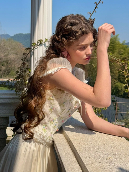 A model leans against a balcony railing, wearing a floral lace corset top with sheer puff sleeves and pastel floral embroidery, styled with a cream flowy skirt, capturing a dreamy, vintage-inspired aesthetic.