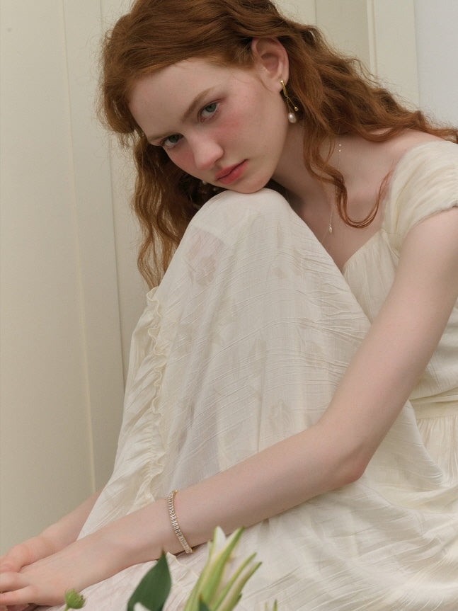 A young woman with red hair gazes forward with a pensive expression, sitting with her knees drawn up, wearing an ivory ruffled dress in a minimalistic indoor setting.