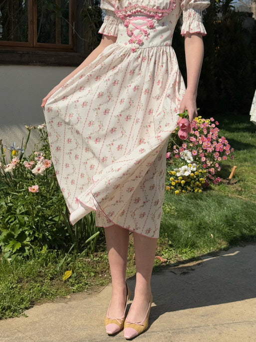 A close-up view of a woman wearing a cream cotton dress with delicate pink floral patterns and lace-up ribbon details, standing in a blooming garden while holding pink tulips.