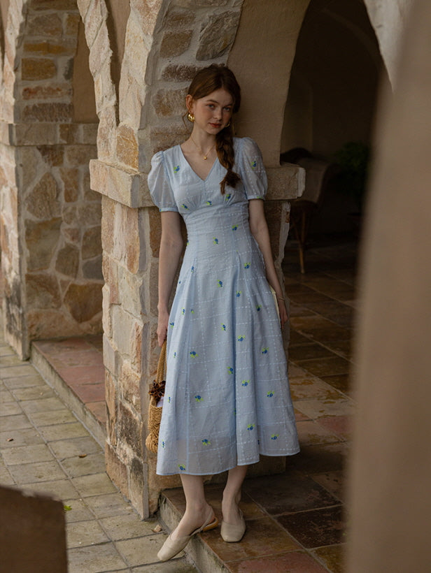 A woman in a pastel blue floral midi dress stands in a stone archway, holding a woven basket bag, exuding a romantic vintage-inspired summer aesthetic.
