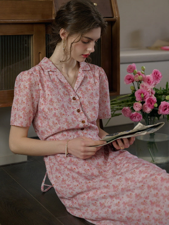 A woman in a vintage-inspired pink floral dress sitting casually on the floor beside a vase of pink flowers, evoking a soft and romantic cottagecore mood.