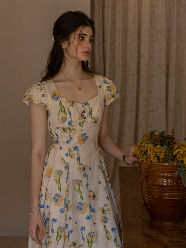 The model stands gracefully by a wooden dining table, holding a woven basket bag and a ripe peach in her hands. Her cream floral dress with blue and yellow flowers flows elegantly, capturing a dreamy, cottagecore atmosphere with classical bust sculptures and rustic candle holders in the background.