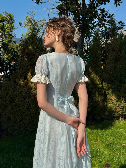 Back view of a woman in a light blue puff-sleeve dress with a tied waist ribbon and lace-trimmed sleeves, standing in a sunlit garden with her hands behind her back.