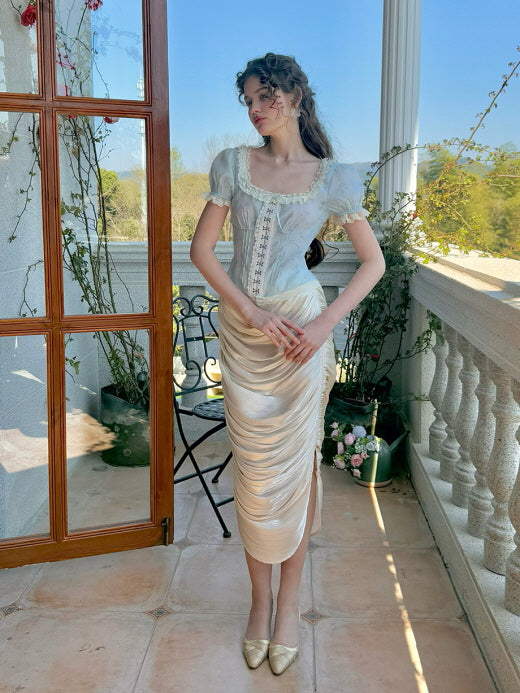 A model poses elegantly in a pastel blue corset top with lace detailing and puff sleeves, styled with a ruched satin midi skirt, creating a soft, vintage-inspired look against a garden backdrop.