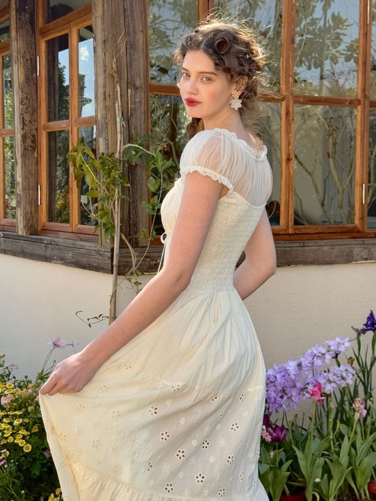 A woman wearing a romantic ivory cottagecore dress with a sheer lace neckline and embroidered floral details poses in front of a rustic wooden house, surrounded by blooming garden flowers, embodying a soft princesscore style.