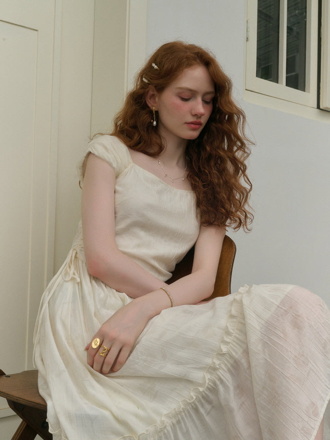 A young woman with red hair sits gracefully on a wooden chair, wearing an ivory vintage-style dress with a gathered waist and ruffle hem, in a softly lit room.