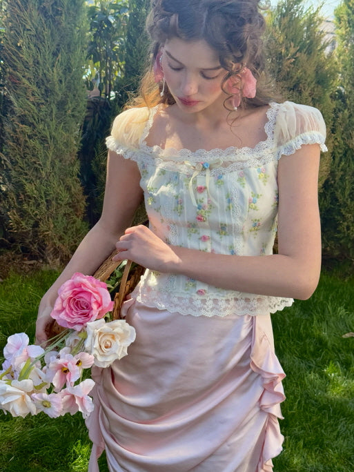 A model holding a woven basket filled with pink and cream roses, wearing a floral lace corset top with sheer puff sleeves and a pink ruched satin skirt, creating a soft, fairytale-inspired look in a garden setting.