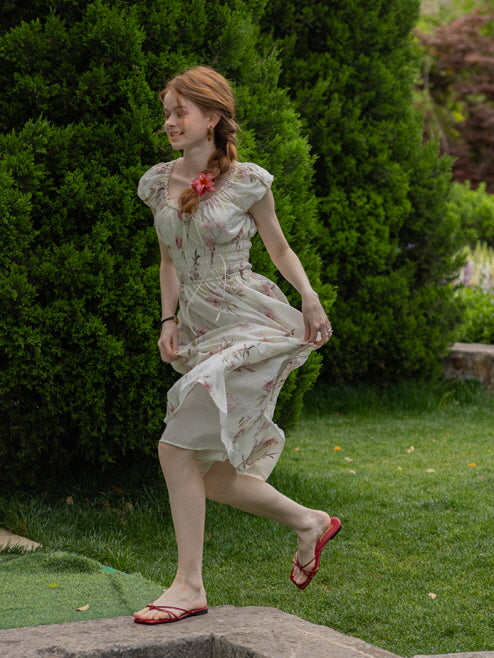 A woman playfully walking through the grass in a floral cottagecore dress with flowing fabric and red sandals. Whimsical, fairytale-inspired style.