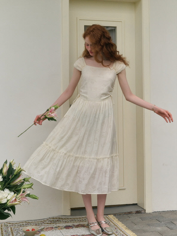 A young woman with red curly hair stands gracefully in front of a cream-colored door, wearing a soft ivory vintage-inspired dress with a ruffled hem, holding a single pink lily flower