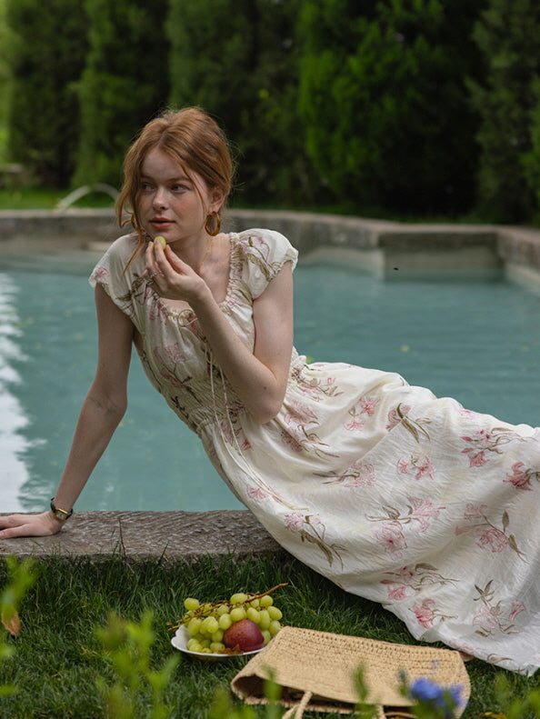 A woman lounging by the water, wearing a floral cottagecore dress with a ruffled neckline and smocked waist. Relaxed, dreamy summer aesthetic.