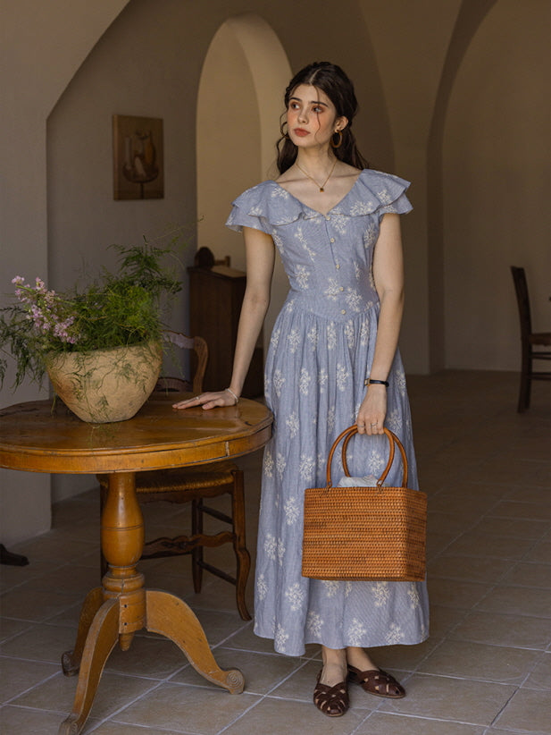 The model stands elegantly beside a rustic wooden table, holding a woven rattan bag in one hand. Her light blue floral dress with a ruffled neckline and button-down front flows gracefully, capturing a classic, vintage-inspired look in a warm, cottage-style room.