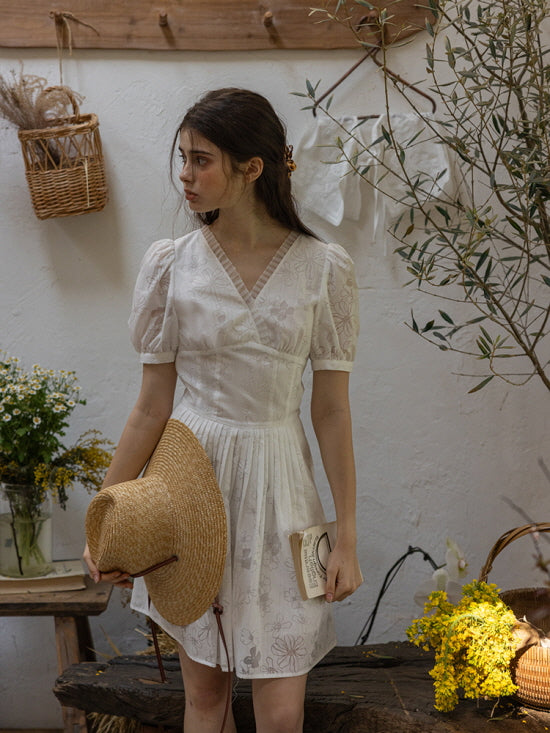 Young woman in a white floral embroidered short dress with puff sleeves and a pleated skirt, holding a straw hat and a small book in a quaint, cottage-style garden scene with hanging linens and scattered yellow flowers.