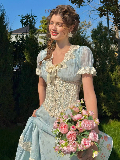 A woman wearing a vintage light blue dress with puff sleeves, lace trim, and a decorative corset, holding a bouquet of pink roses while smiling in a sunlit garden.