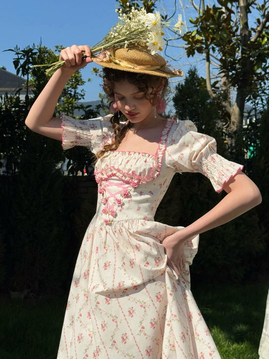 Wearing a vintage-inspired cotton dress with a corset-style pink ribbon detail and puff sleeves, the model poses in a sunny outdoor garden, surrounded by flowers — a perfect look for women who love romantic fairytale style.