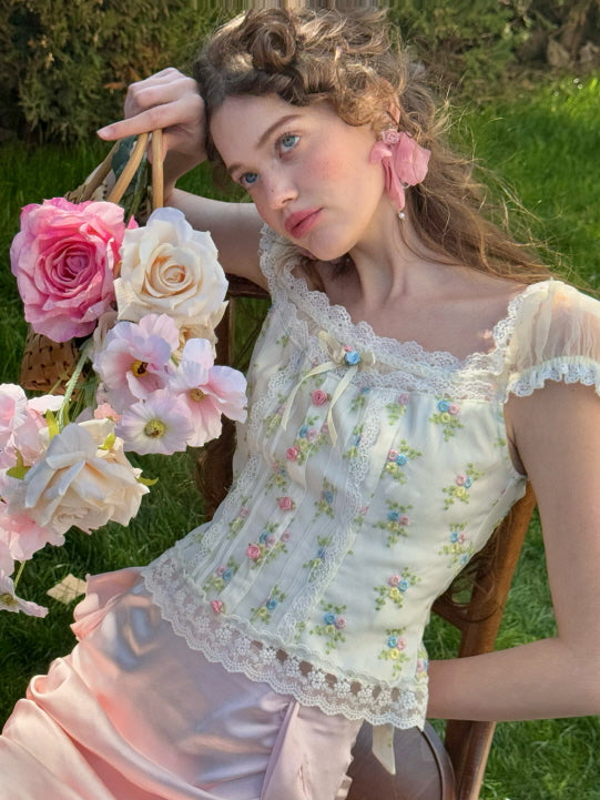 A model reclining on a chair, holding a basket of roses while wearing a floral lace corset top with pastel floral embroidery and a pink satin ruched skirt, exuding a dreamy, vintage aesthetic against a lush green background.