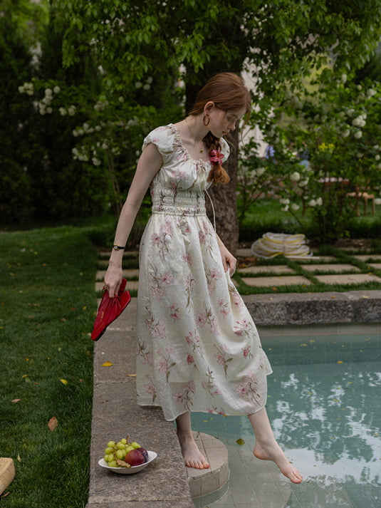 A woman dipping her toes in the water while holding red sandals, wearing a floral cottagecore dress with a smocked waist and ruffled sleeves. Ethereal summer look.