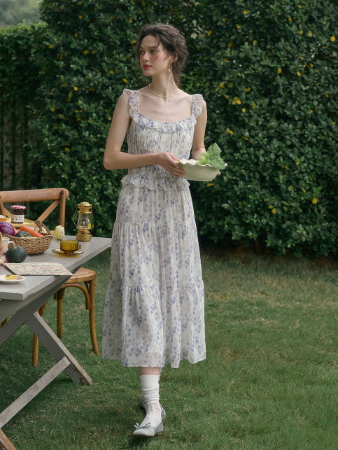 Woman wearing a cottagecore floral outfit with ruffle straps, standing on grass holding a bowl of grapes, surrounded by vintage picnic setting – aesthetic fashion.