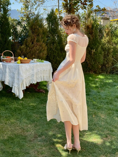 Back view of a woman in an ivory lace-trimmed fairycore dress walking barefoot on the grass, beside a vintage outdoor tea table — romantic, soft vintage look from Romantic Fairytale.