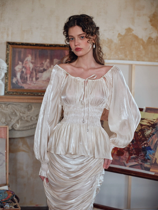 A model wearing a romantic ivory blouse with voluminous bishop sleeves and a ruched waist, standing in front of vintage-style artwork and decor. The soft sheen and peplum silhouette evoke a feminine vintage aesthetic.