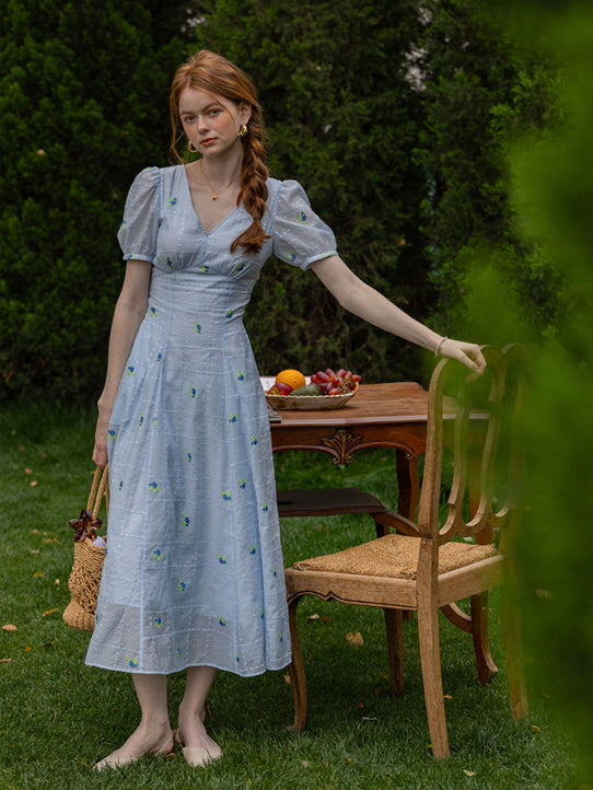 Outdoor scene featuring a woman in a light blue floral midi dress with puff sleeves and a fitted waist, holding a woven basket while standing beside a vintage wooden chair and a table set with fruit, evoking a romantic summer cottagecore vibe.