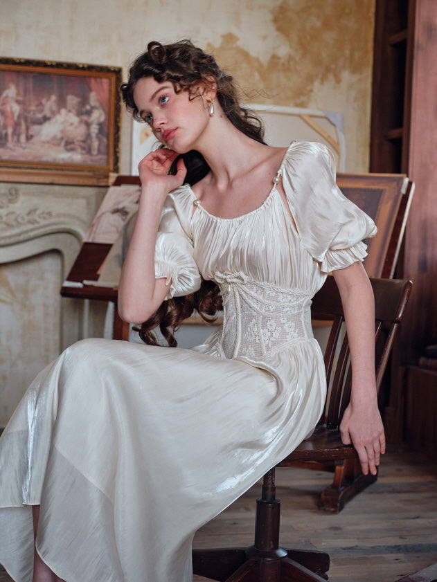 A woman seated thoughtfully in an ivory puff-sleeve dress with pearl details and an embroidered lace corset, surrounded by classical artwork and a softly textured interior.