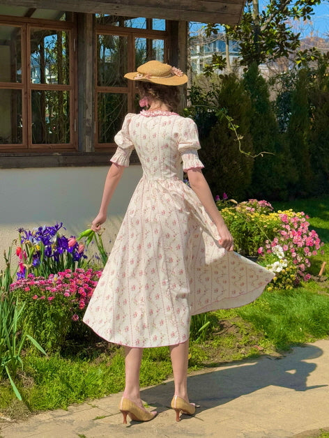 A woman wearing a pink floral cotton dress stands in a blooming garden, her back facing the camera as she gently lifts the skirt, evoking a romantic fairytale atmosphere with a vintage aesthetic.