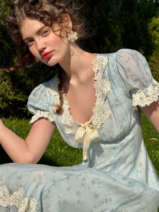 A woman wearing a vintage-inspired light blue dress with puff sleeves, ivory lace trim, and a satin bow, styled with floral earrings and curly hair, sitting in the sunlit garden.