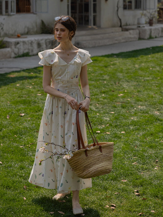 Young woman standing in a cottagecore floral dress with flutter sleeves, holding a woven basket filled with blooming branches in a tranquil garden setting.