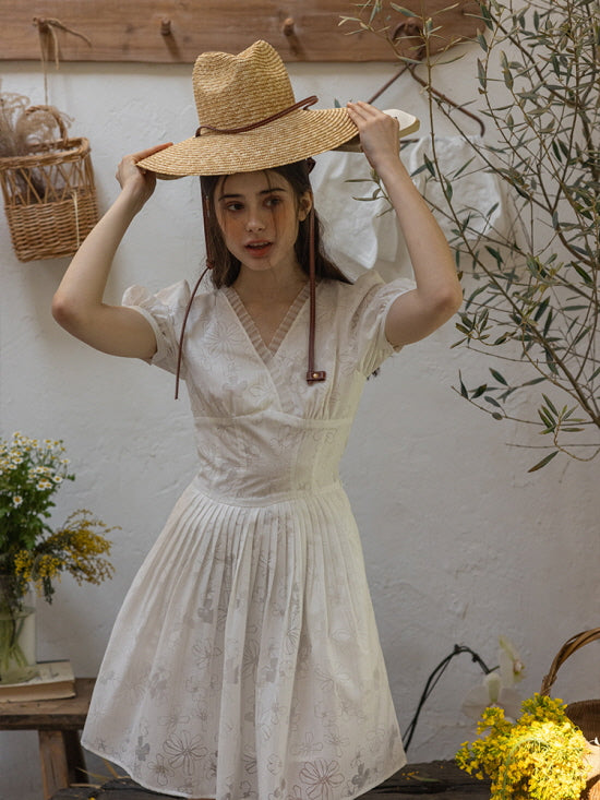 Young woman wearing a white floral embroidered short dress with puff sleeves and a pleated skirt, adjusting a wide-brimmed straw hat in a rustic, cottagecore-inspired garden setting.