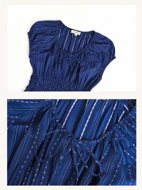 Flat lay of a navy cap-sleeve dress with fine stripes and front tie detail at the neckline. Close-up shows ruched gathering and feminine bow accent on soft, lightweight fabric.