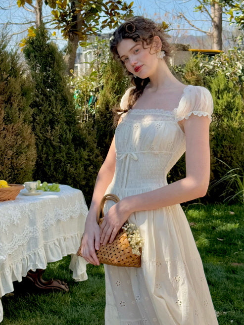Model wearing a romantic cream eyelet midi dress with puff sleeves and lace-trimmed square neckline, standing in a garden with a woven basket – cottagecore and princesscore aesthetic for women.