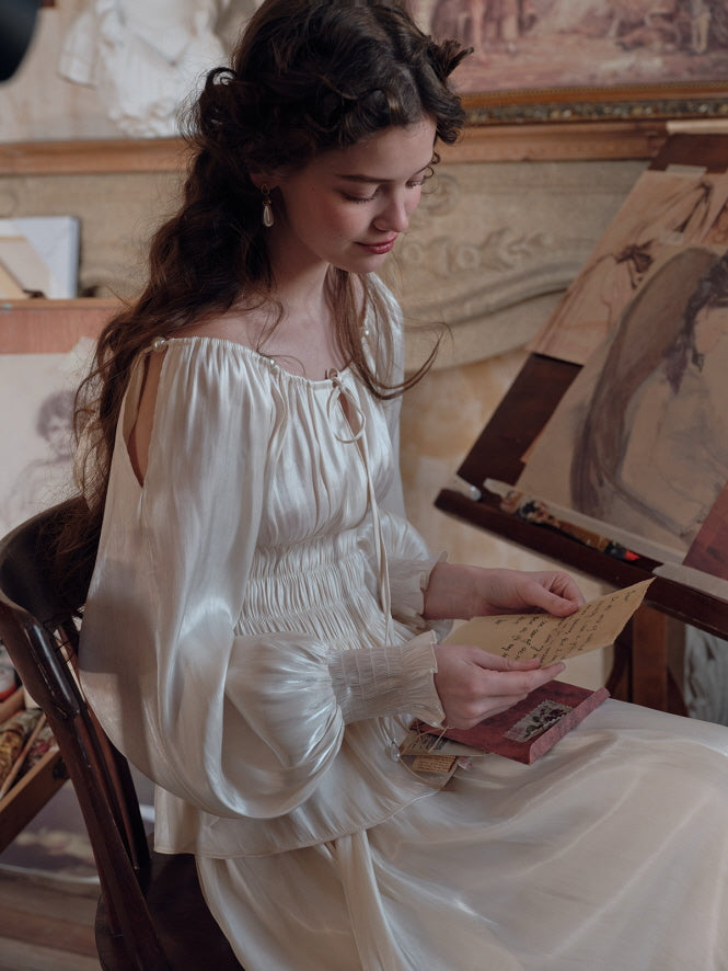 A young woman in an ivory satin blouse with shirred details and balloon sleeves, sitting at an art desk while reading a letter, surrounded by classical sketches and paintings. The setting enhances the dreamy, cottagecore mood.