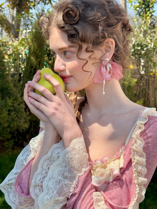 A young woman in a vintage-inspired pink floral dress with lace-trimmed sleeves, holding a green pear close to her face in a dreamy, fairytale-like garden setting.