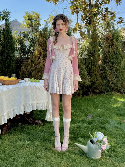 A woman standing in a lush garden, wearing a pink floral mini dress with a matching lace-trimmed cardigan, paired with lace socks and pastel pink heels, capturing a romantic, cottagecore aesthetic.