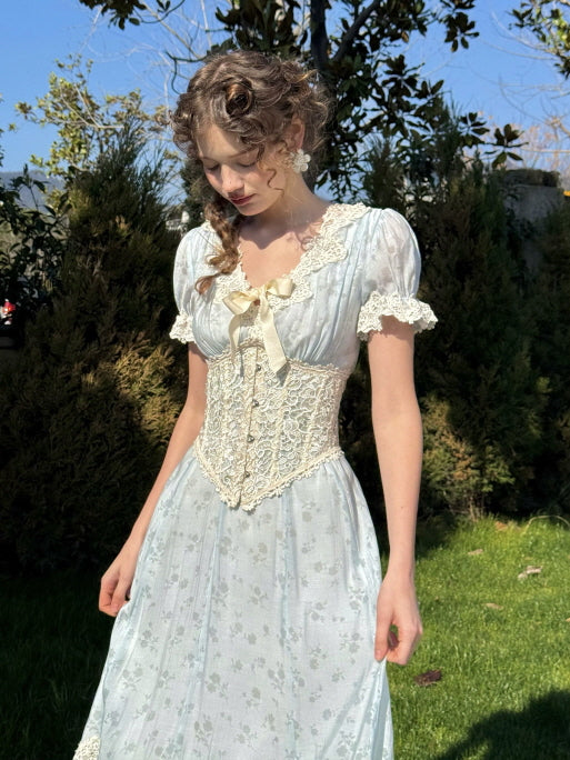 A woman standing in a sunlit garden wearing a vintage-style light blue dress with puff sleeves, lace trim, and a decorative ivory lace corset, styled with floral earrings and braided hair.