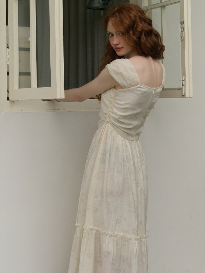 A woman with wavy red hair wearing a soft ivory dress stands by an open window, looking back over her shoulder with a gentle smile. The dress features ruched detailing at the waist and a square neckline, creating a romantic and vintage-inspired silhouette.