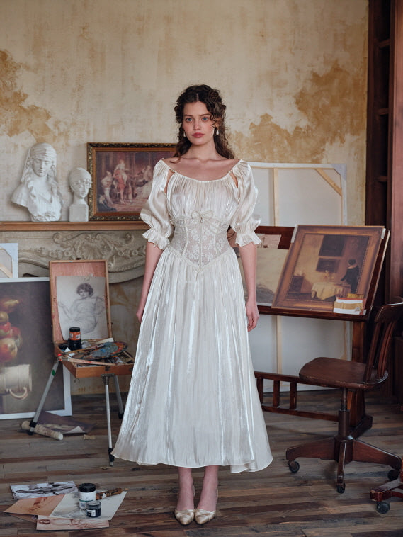 A woman standing in an artist’s studio wearing a vintage-inspired ivory dress with puff sleeves and a pearl-accented lace corset, surrounded by classical sculptures and paintings.