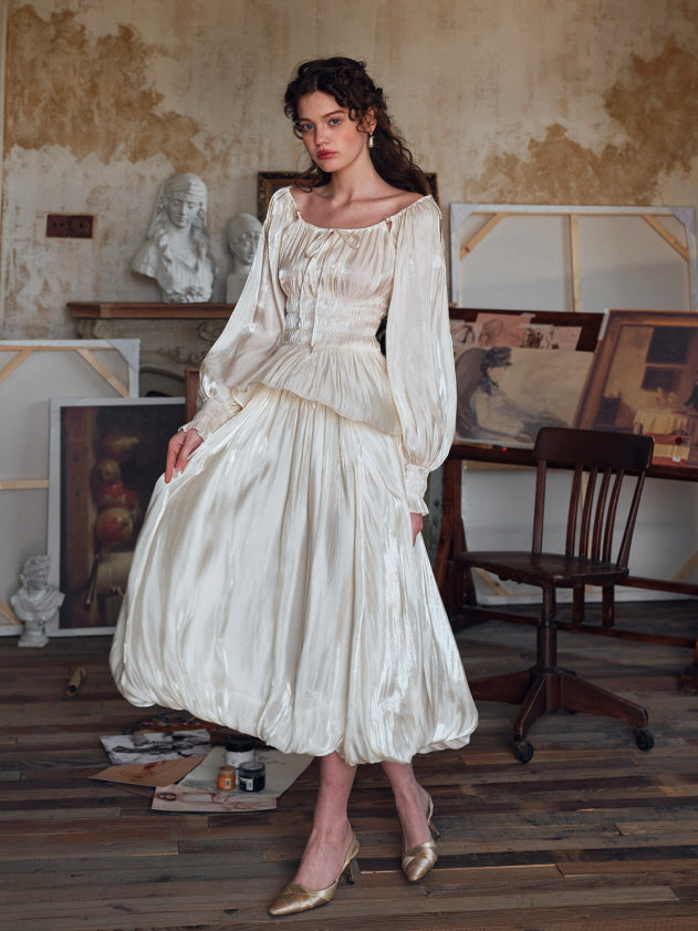 Model wearing a puff-sleeve vintage-inspired ivory outfit with a bubble hem skirt, posing gracefully in an art studio surrounded by paintings and wooden flooring – romantic aesthetic.