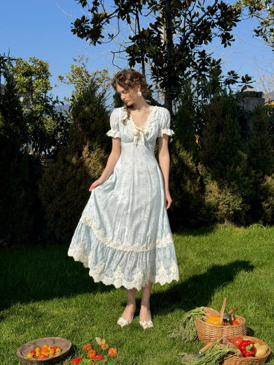 A woman standing in a sunny garden wearing a light blue vintage dress with lace trim and puff sleeves, gently lifting the hem of her dress while surrounded by vegetables and a woven basket on the grass.