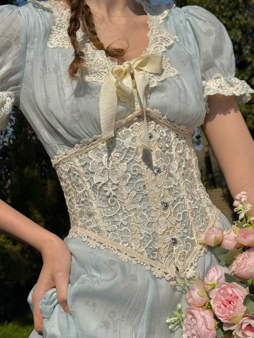 Close-up of a light blue dress featuring intricate ivory lace corset detailing, puff sleeves, and a satin bow, with the model holding a bouquet of pink flowers in a fairytale-inspired garden setting.