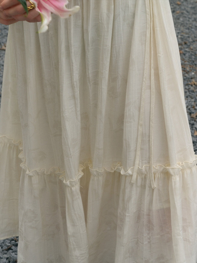 A close-up of the skirt of a light ivory dress featuring soft gathered tiers and subtle floral patterns, with a hand holding a pink lily flower slightly entering the frame.